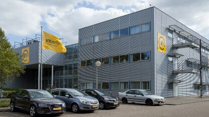 Exterior view of the multi-story Intertek Geleen polymer testing laboratory which has grey walls with yellow signage and flags