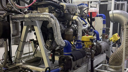 A close side view of an engine installed on a test bed, with multiple cables, sensors, insulated pipes, and coolant hoses connected for measurement. Industrial equipment, ventilation ducting, and control units surround the setup inside the test cell.